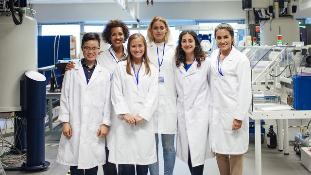 A group of six female scientists stand together in a lab wearing lab coats.